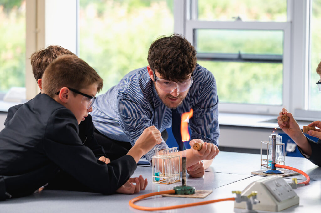 A teacher wearing glasses lighting a bunsen burner with two pupils