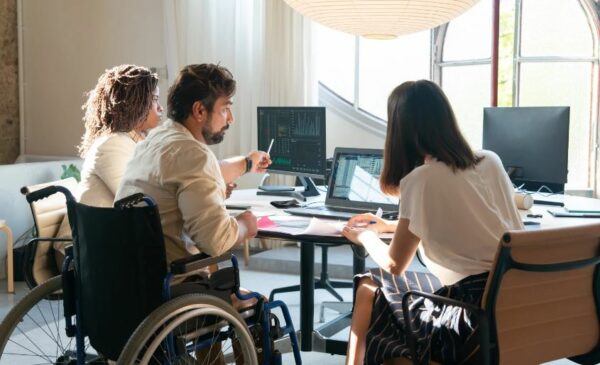 Three people, one who is in a wheelchair, sitting at a desk and having a conversation.