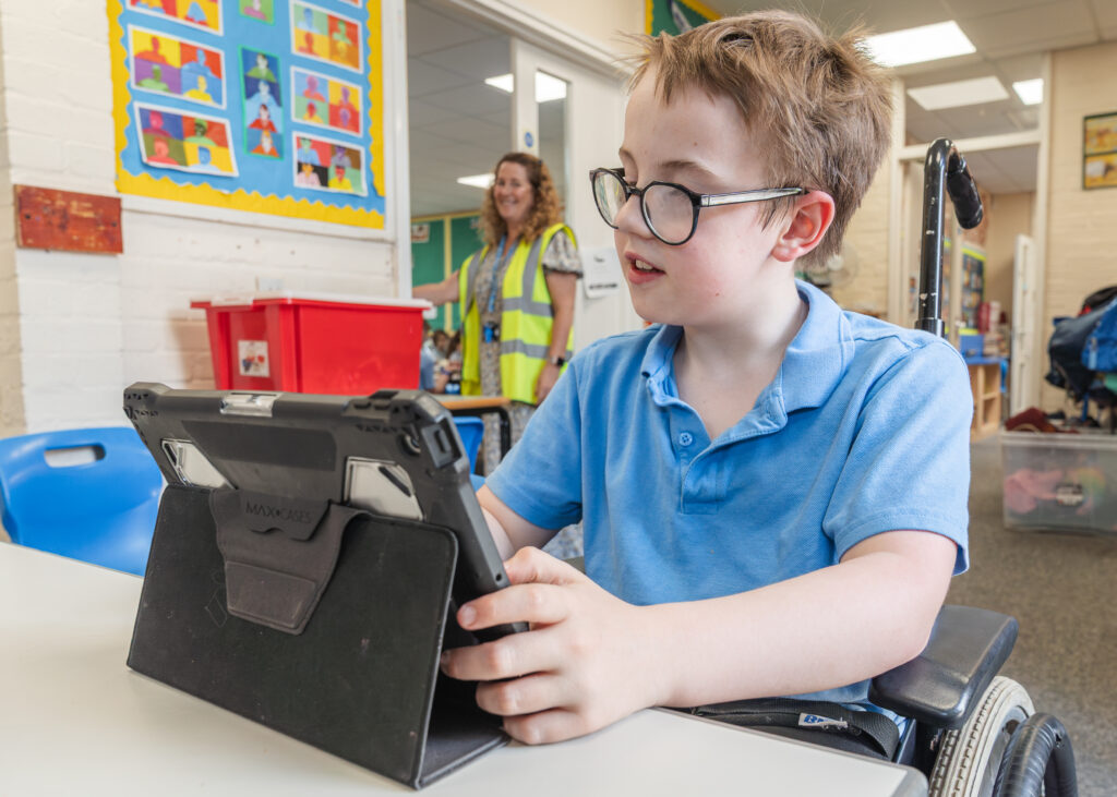 A pupil in a wheelchair is sitting at a desk and holding an iPad. He is wearing glasses and a blue polo shirt.