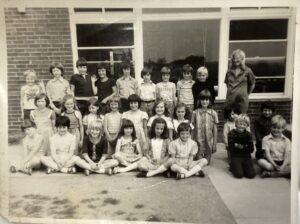 A black and white photograph of a class of school children sitting in a row and smiling at the camera.