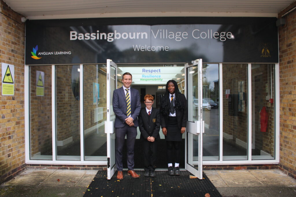 A man stands infront of an entrance with a sign that reads Bassingbourn Village College with two pupils