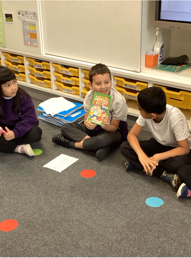 A young boy holding up a green child's book and showing it to two other pupils sitting down on the floor