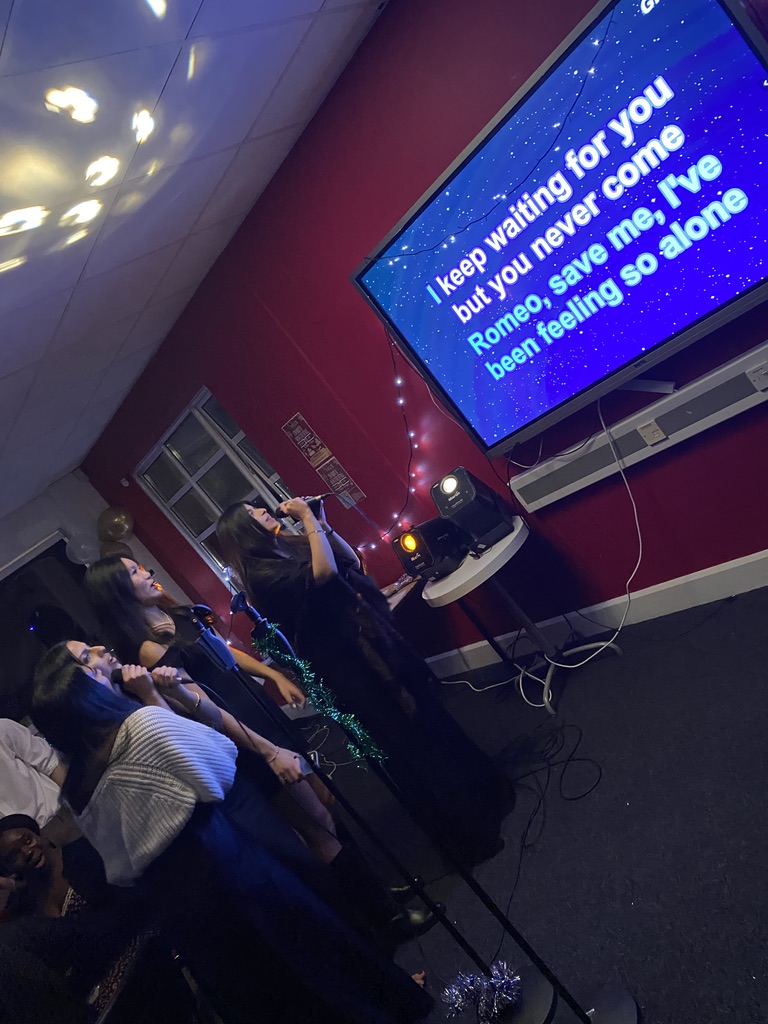 In a dimly lit room, three people are facing towards a TV screen which contains lyrics to Taylor Swift's song Love Story with some of the wording highlighted in bold.