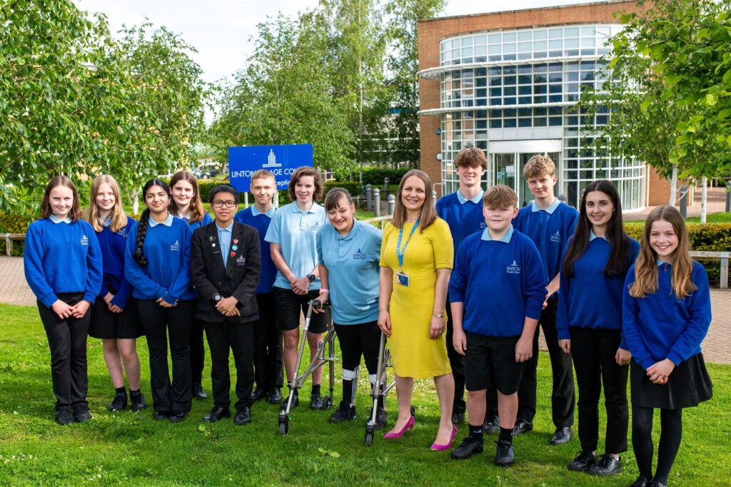 A large group of pupils wearing blue school jumpers and black trousers and standing with a lady in a yellow dress. One pupil is using standing using a wheelchair support device. They are all smiling at the camera