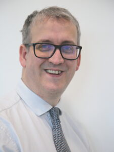 A white man wearing glasses who is wearing a white shirt and tie is smiling at a camera.