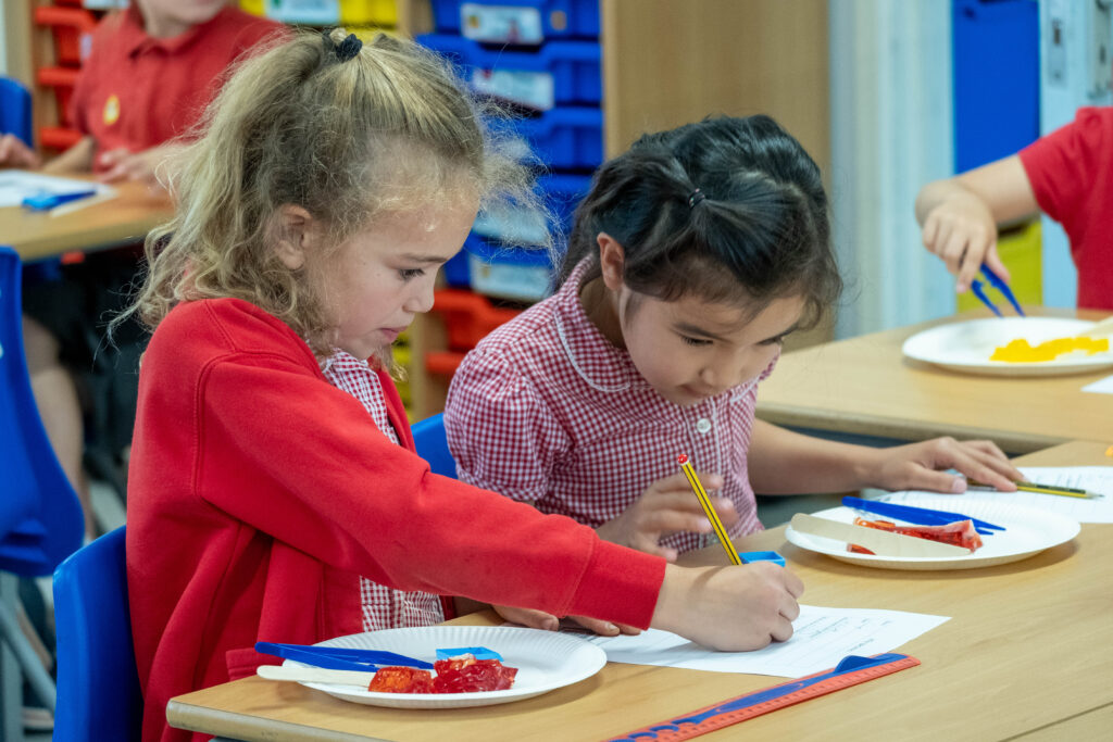 Two pupils, sitting at a desk, are drawing on a paper plate. One child is wearing a red cardigan, the other is wearing a red and white gingham dress. Both are looking at their drawings. 