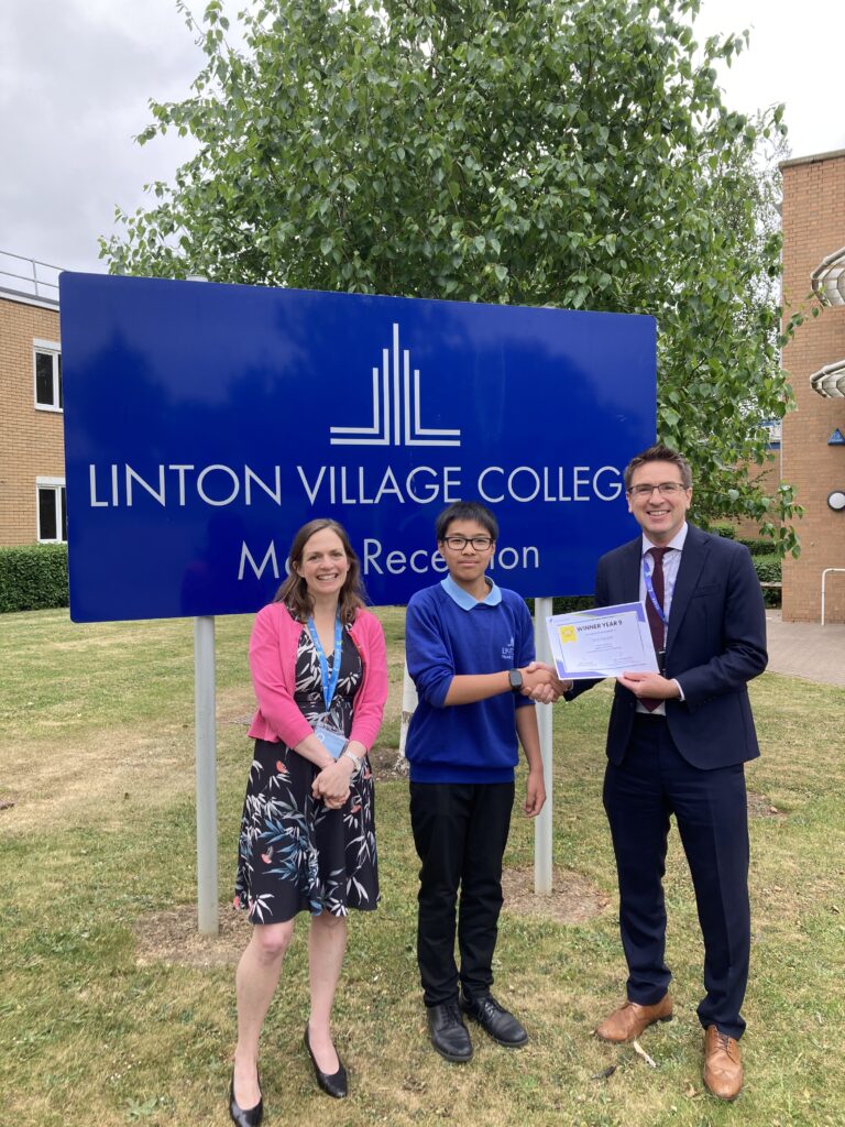Three people are standing in front of a sign that reads 'Linton Village College'. The woman on the left is wearing a pink cardigan with a floral dress. The pupil standing in the middle is wearing a blue jumper and black trousers. The man on the right is wearing a black suit, and is holding a certificate and shaking the pupils hand.