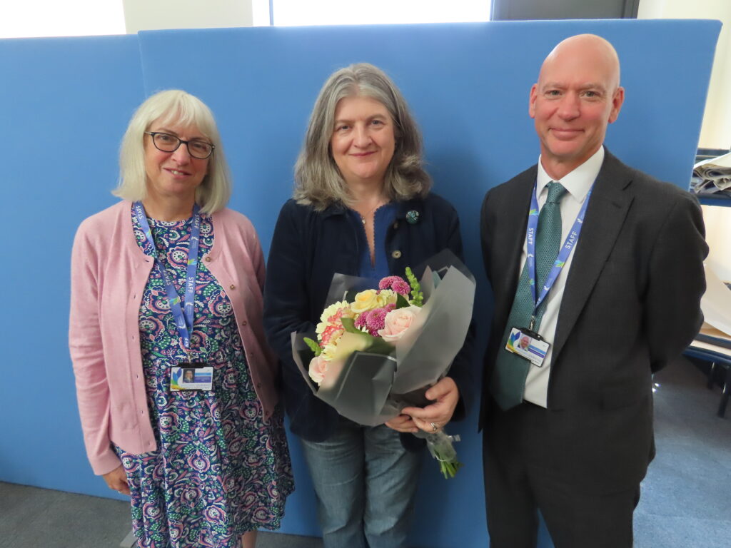 Three people stand in front of a blue board. On the left, a lady with short, white hair is wearing a colourful dress and pink cardigan. In the middle, a lady with long grey hair is wearing a black cardigan with jeans and is holding a bunch of flowers. On the right, a man is wearing a suit with a blue tie. All are smiling at the camera. 