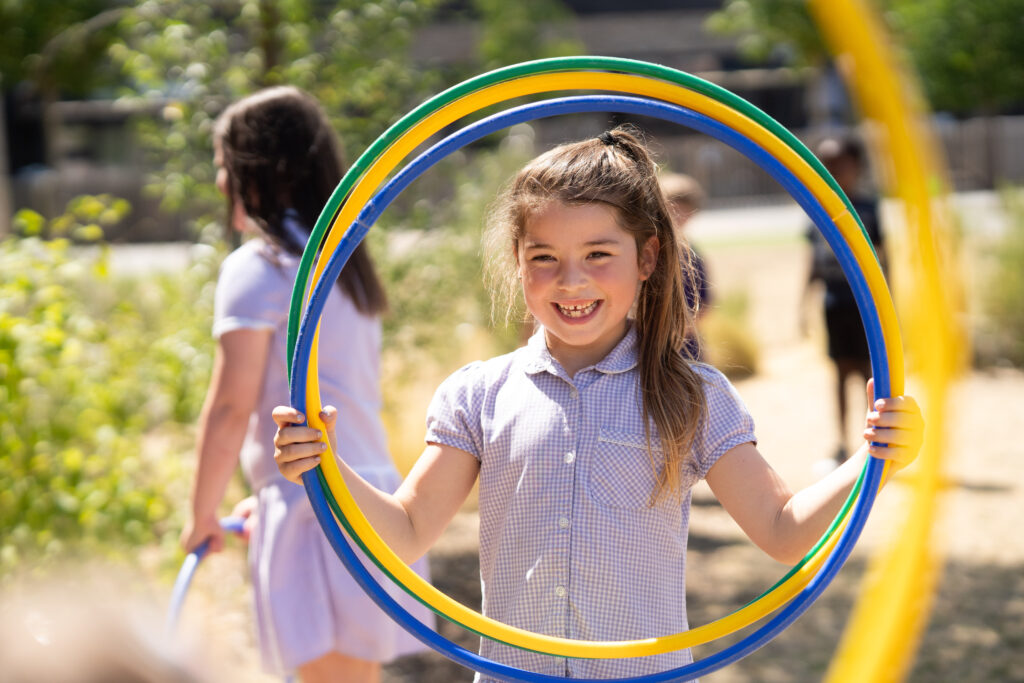 A pupil wearing a purple checkered dress is holding up three hula hoops to her head and smiling to the camera within the middle circle.