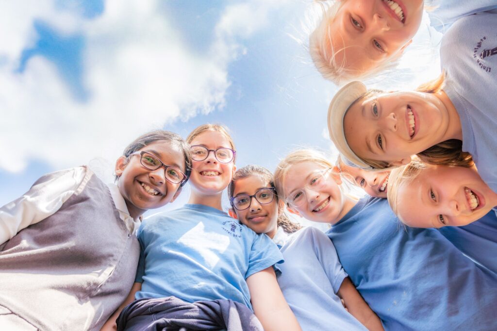 six pupils are smiling at a camera which has been placed on the ground. They are all wearing a mixture of blue tops and blue dresses.