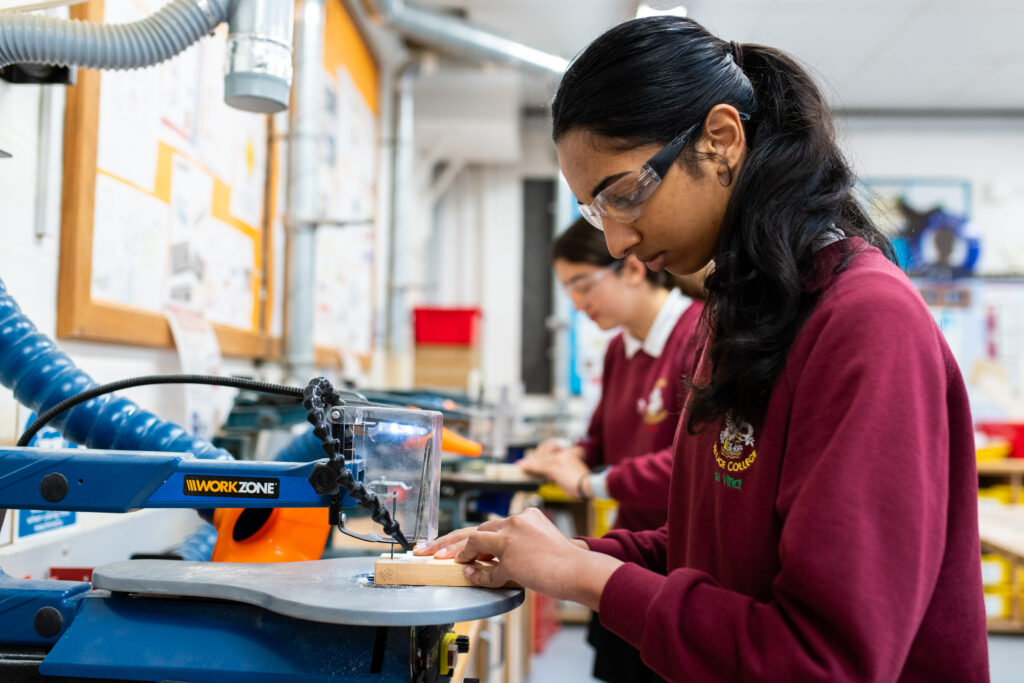 Two pupils, both wearing burgundy jumpers and protective eyewear, are working on a machine which is cutting a small piece of wood.