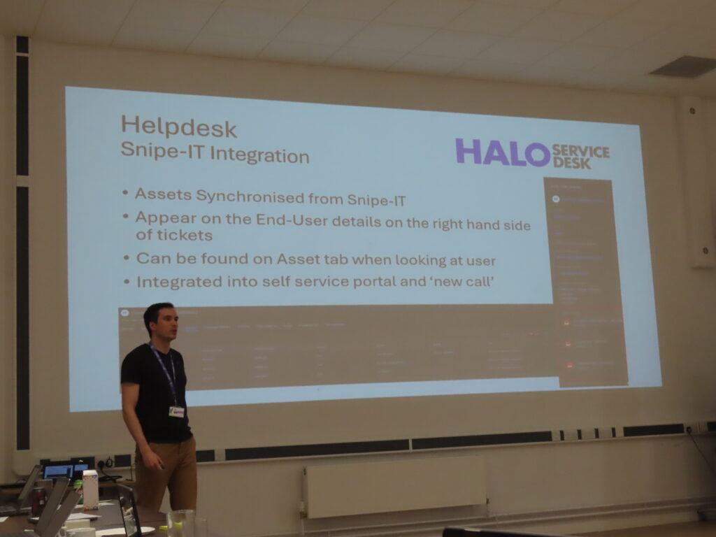 A man wearing a black t-shirt and beige shorts is standing in front of a large white projector screen. He is presenting to a group of people who can not be seen on the shot.