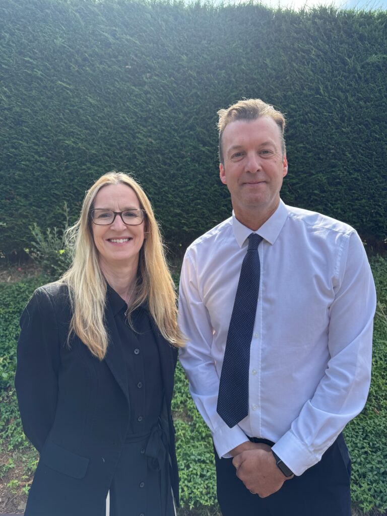 A man and a woman are standing next to each other. The man is wearing a white shirt and tie, and the woman is wearing a navy top and suit jacket. 