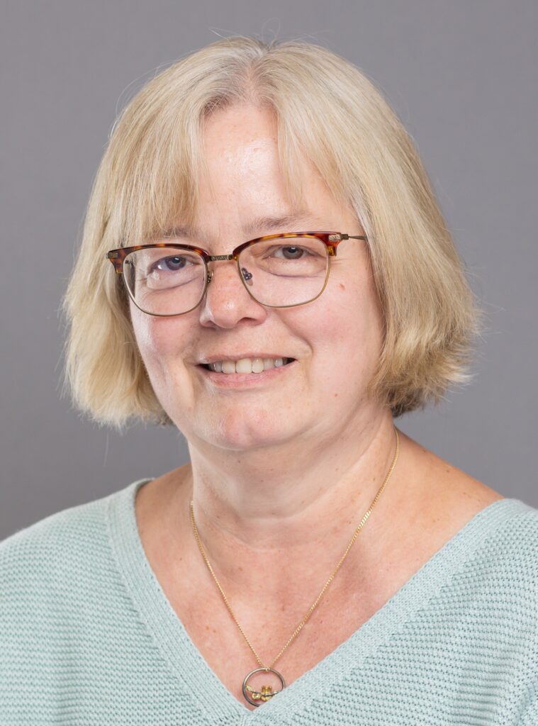 A headshot of a white woman with short, blonde hair and glasses who is smiling at the camera. She is wearing a blue, v-neck top.