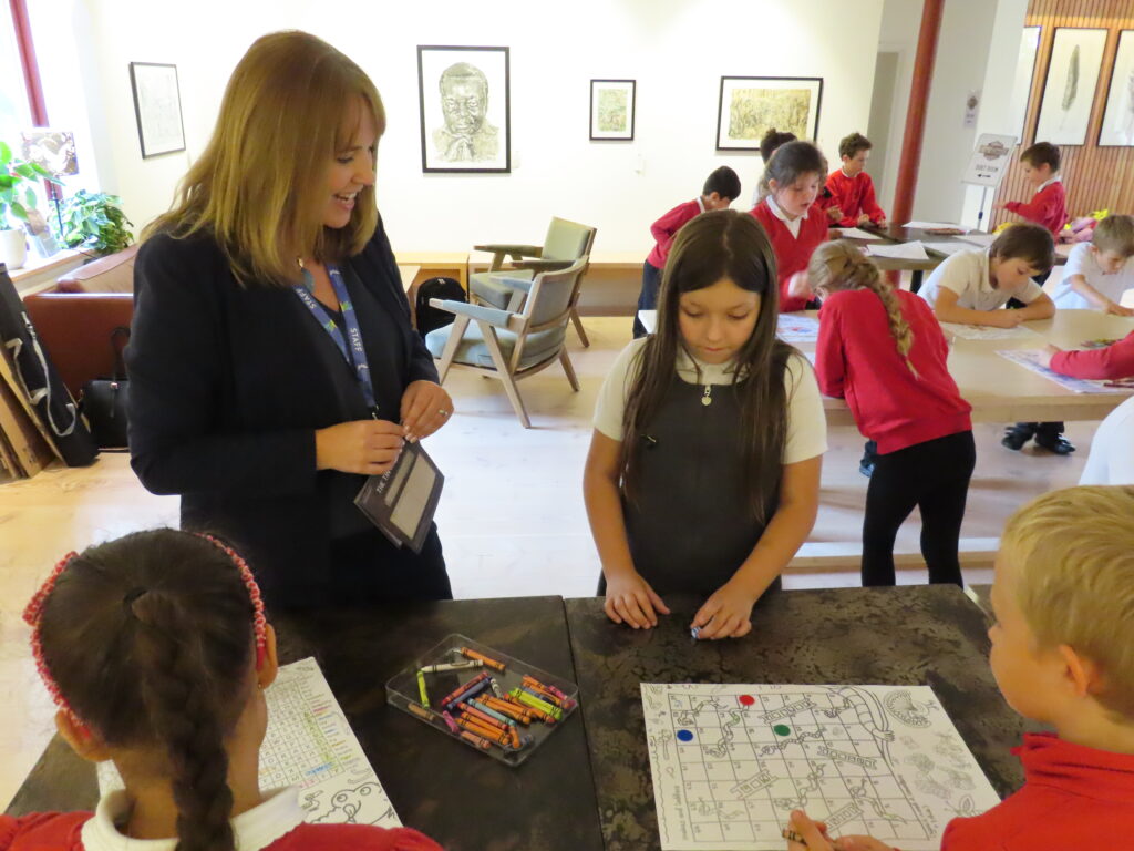 A women with long brown hair and wearing a black suit jacket is standing at a desk. She is watching two pupils playing snakes and ladders.