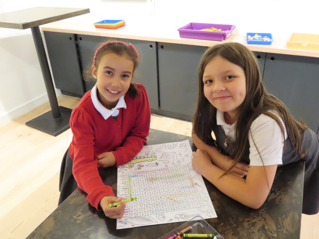 Two pupils are standing at a desk. One is wearing a red jumper and pink headband. The other is wearing a white polo shirt underneath a grey dress. They are completing a wordsearch. They are smiling at the camera.