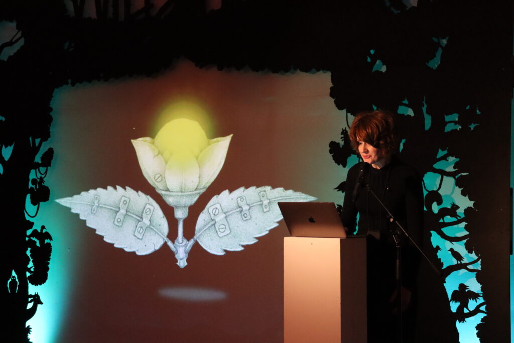 A women with short, brown hair and wearing a long black dress is standing on a podium with has a laptop placed on top of it. Behind her is screen with a flower made out of metal and a bright, yellow bulb in the middle.