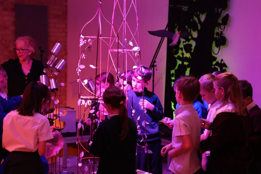 A woman with short, blonde hair is standing with a number of children dressed in school uniform. They are circled around a tall, thin metal structure. They are tying small paper leaves to the structure in a darkened room.