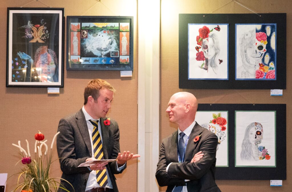 Two men are standing in front of artwork placed on a wall. They are both wearing black suits with red poppies attached to their left shoulder. 