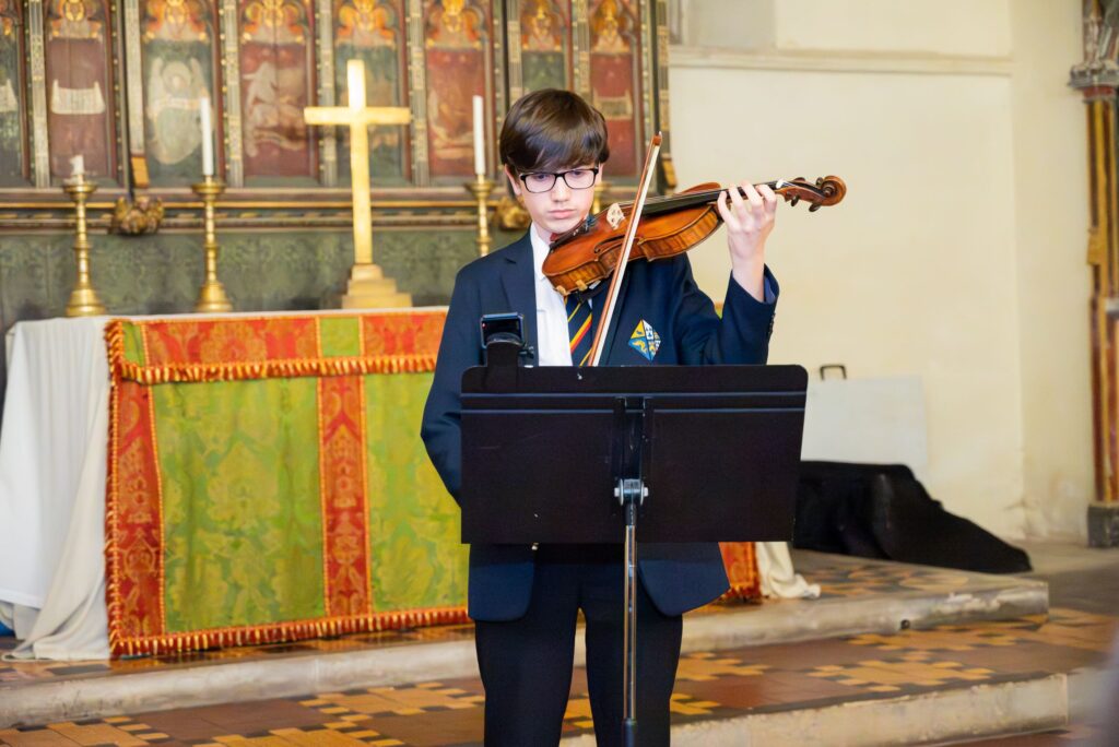 A boy wearing school uniform is playing the violin. He is standing behind a black music stand.