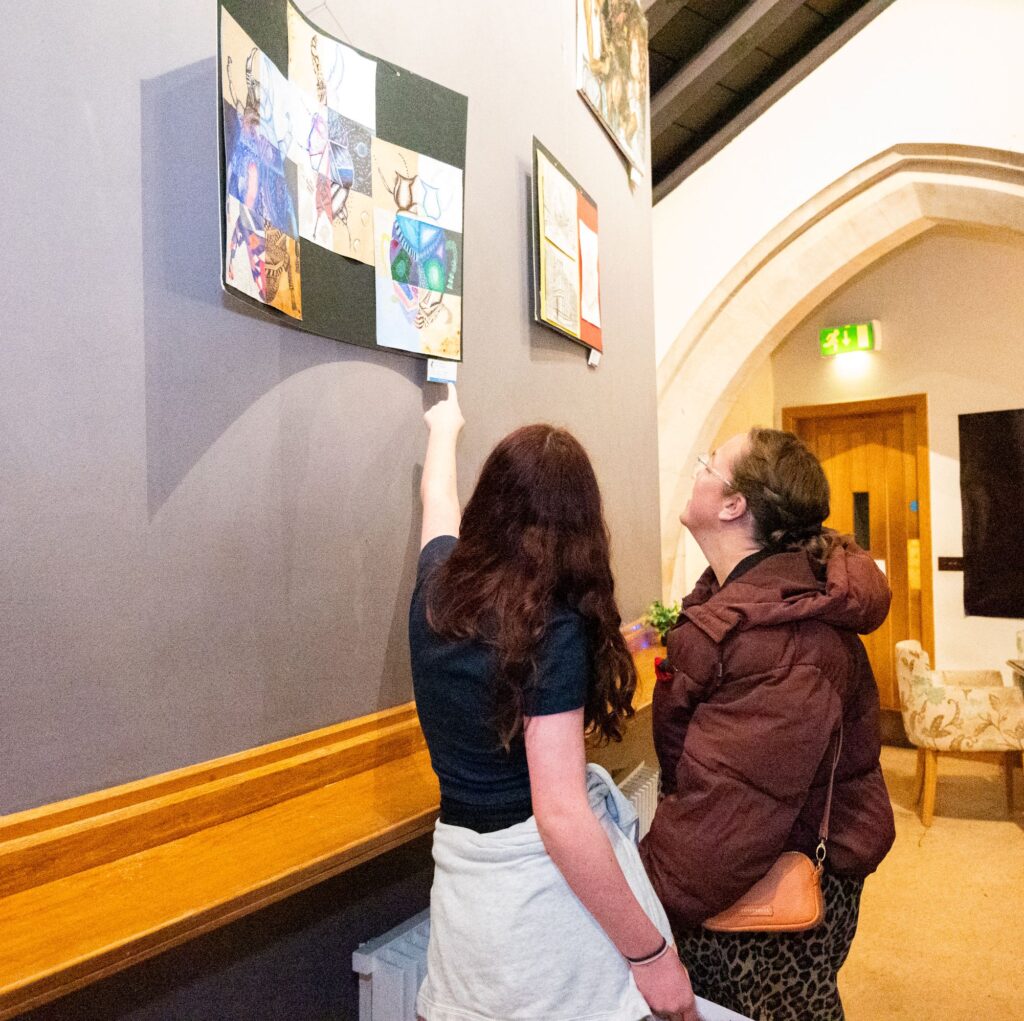 Two people are standing next to a tall wall which contains multiple artworks that has been hung on the wall.