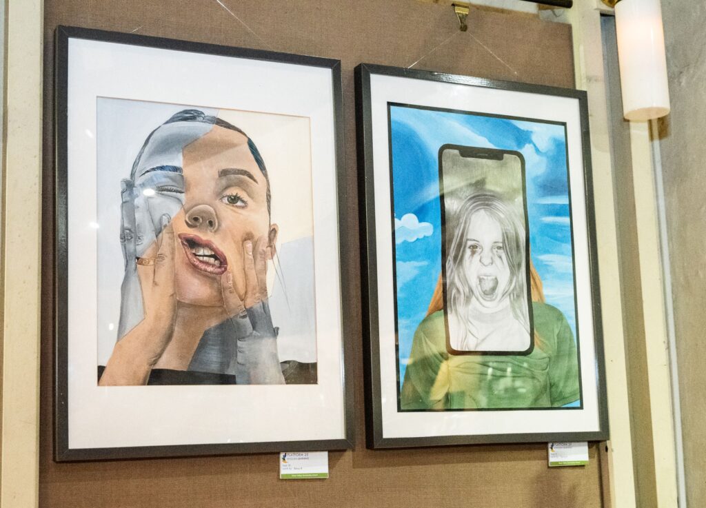 Two artworks are hanging on a wall. One shows a young girl's face which she is pulling down. The other artwork shows a young girl screaming within a mobile phone screen.
