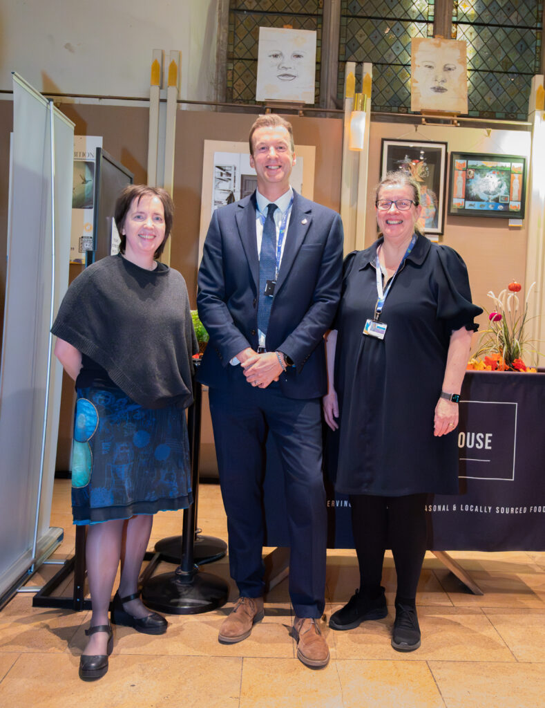 A man is standing between two women smiling for a picture. The woman on the left is wearing a poncho and colourful skirt, the man is wearing a navy suit and blue tie, and the woman on the right is wearing a navy dress. 