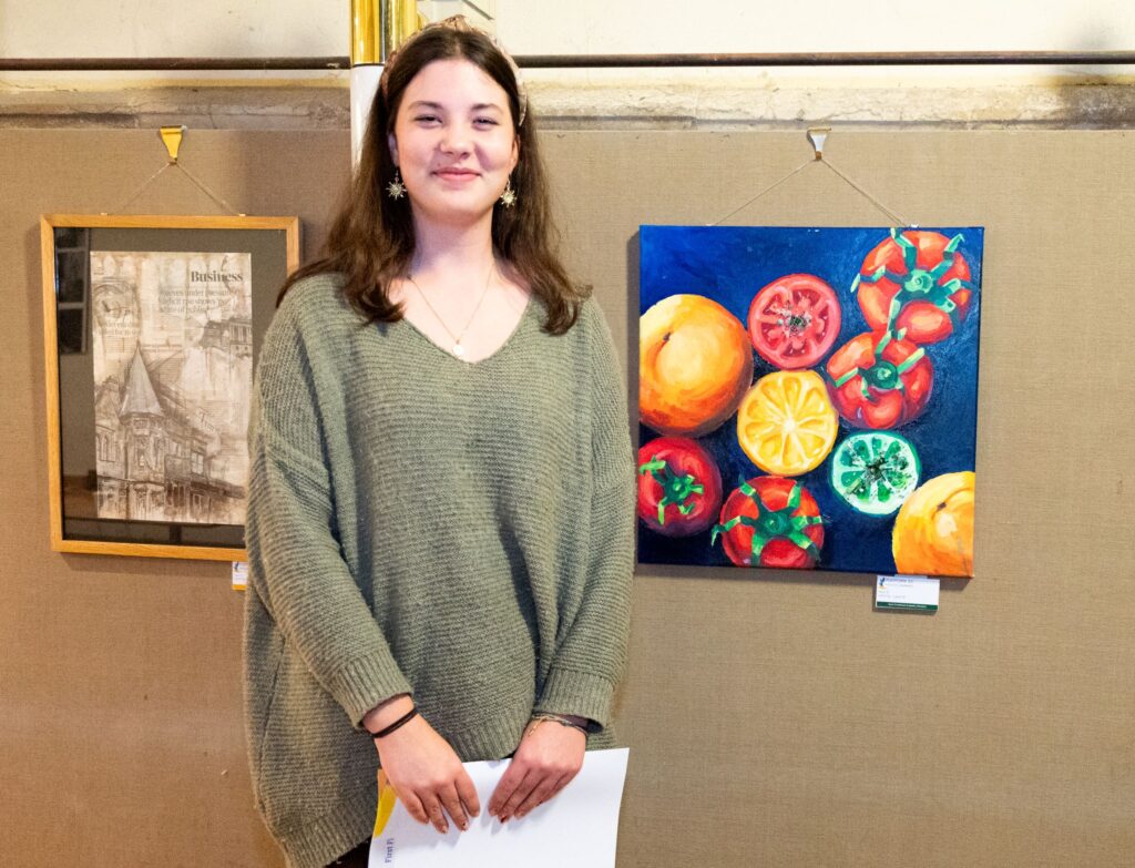 A woman who is wearing a green jumper is standing in front of a piece of artwork and smiling at the camera. The artwork is tomatoes painted in green, yellow and red.