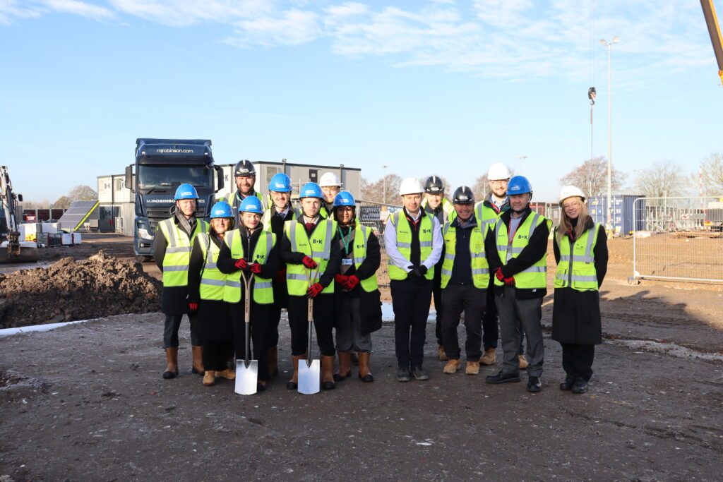 A group of people are standing on a construction site. Two pupils are holding spades. All are wearing yellow reflective jackets and blue and white hard helmets.
