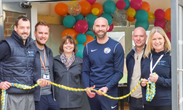 A group of people stood together outside the entrance of the new sports centre. They are stood in front of a wall of celebratory balloons, and the person in the middle of the group is cutting open a ribbon.