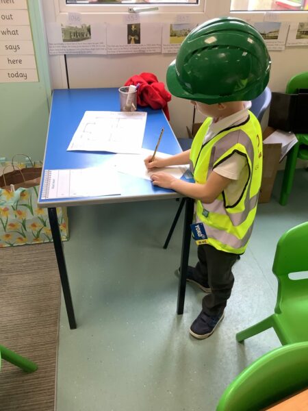 Young child wearing a hard helmet and high-vis jacket writing on a piece of paper.