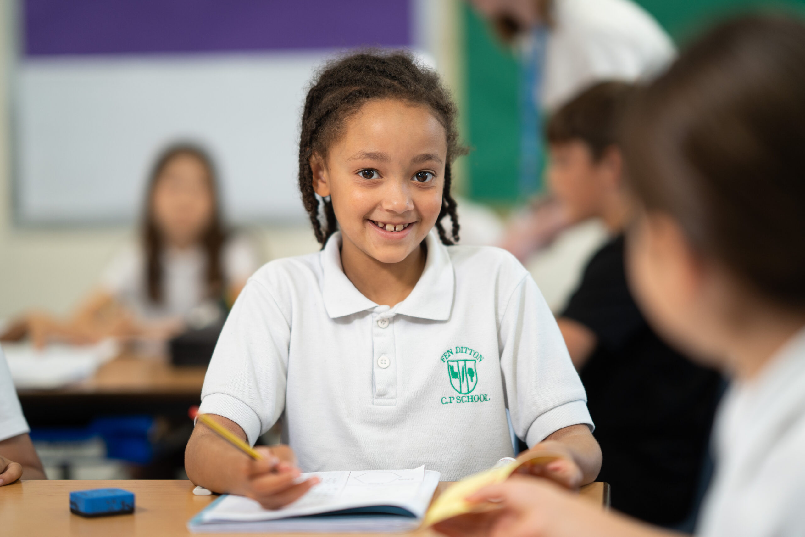 A young girl wearing a white polo shirt is smiling at the camera. She is sitting at a desk and writing in a blue book.