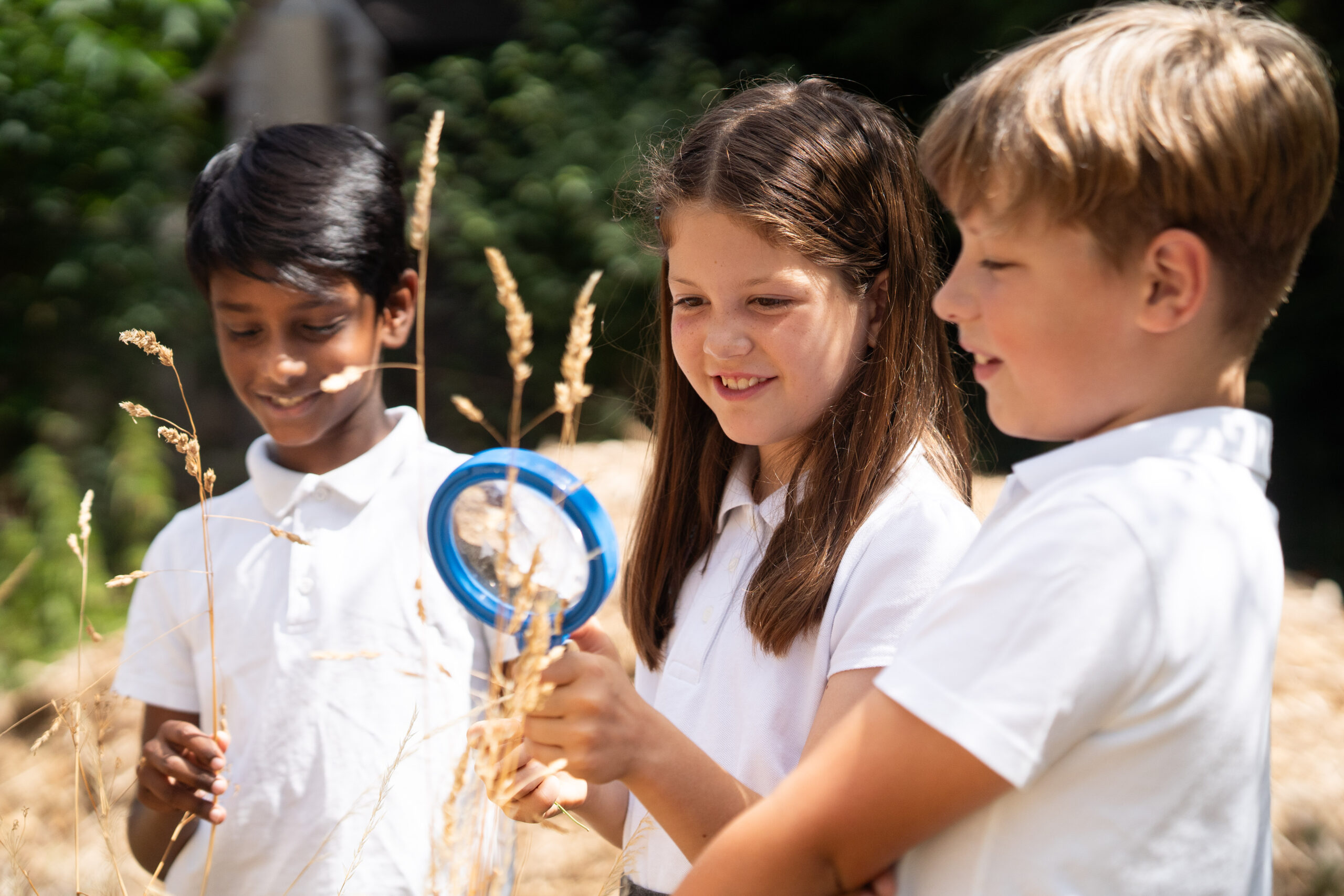 Three pupils are outside in a field. They are all wearing white polos. One is holding a small blue bat and smiling at it.