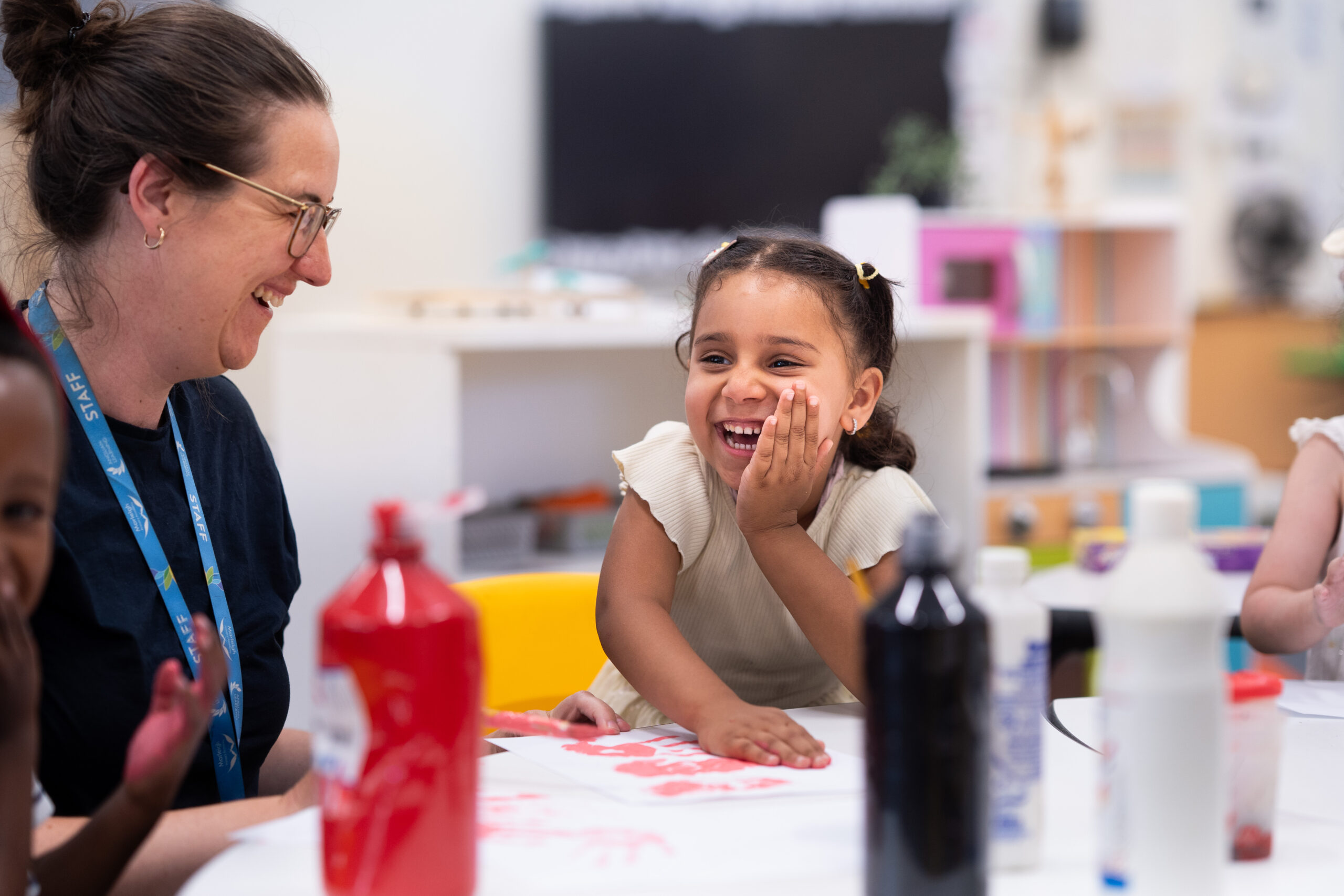 A teacher and pupil are sitting at the table. The table has bottles of paint and a white piece of paper with a painting half finished. The teacher and pupil are looking at each other and smiling.