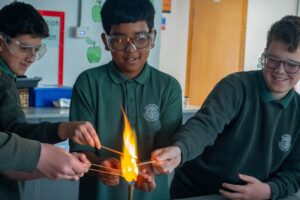 Three pupils are standing in front of a lit Bunsen burner. They are placing rods of metal into the Bunsen burner.
