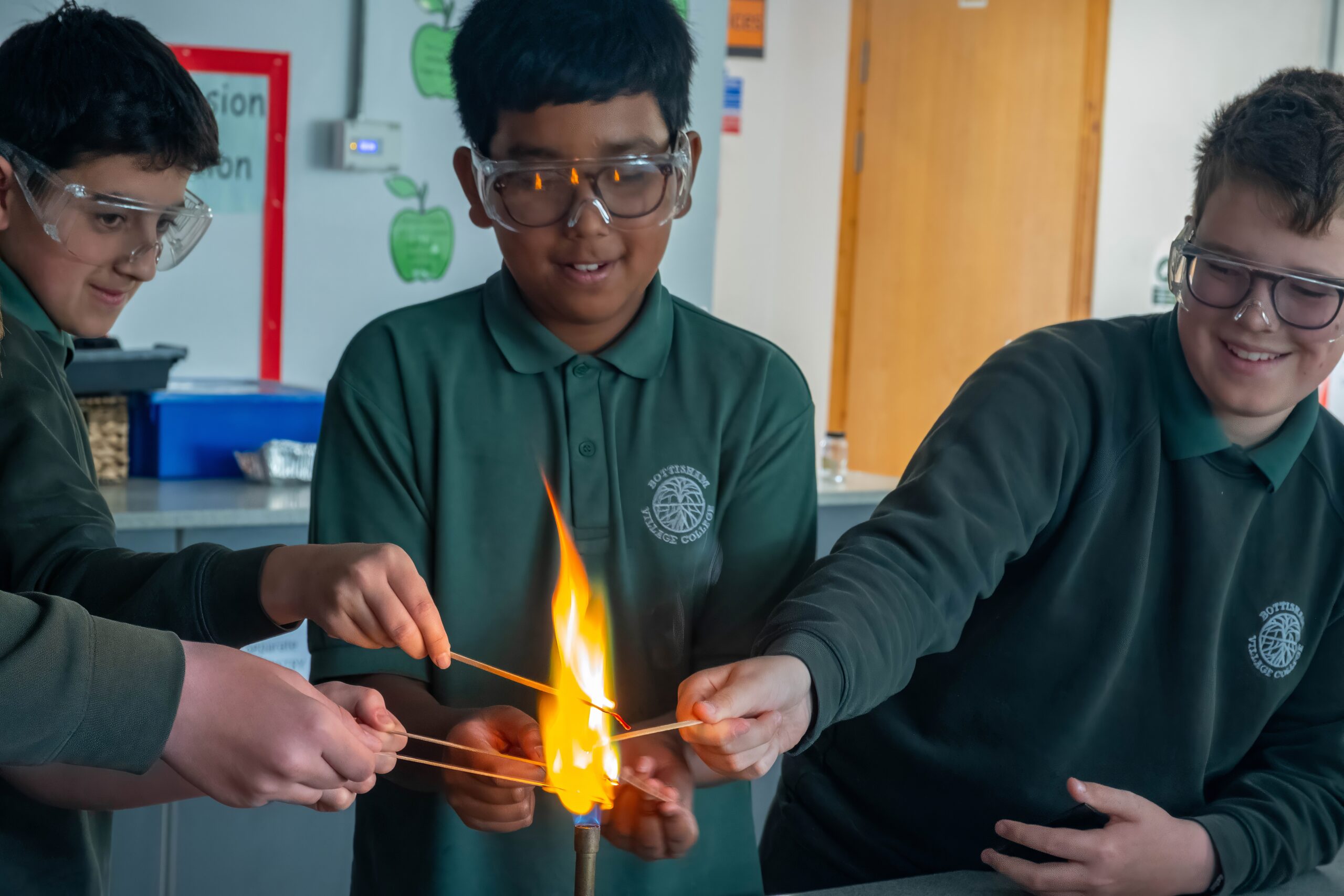 Three pupils are standing in front of a lit Bunsen burner. They are placing rods of metal into the Bunsen burner.