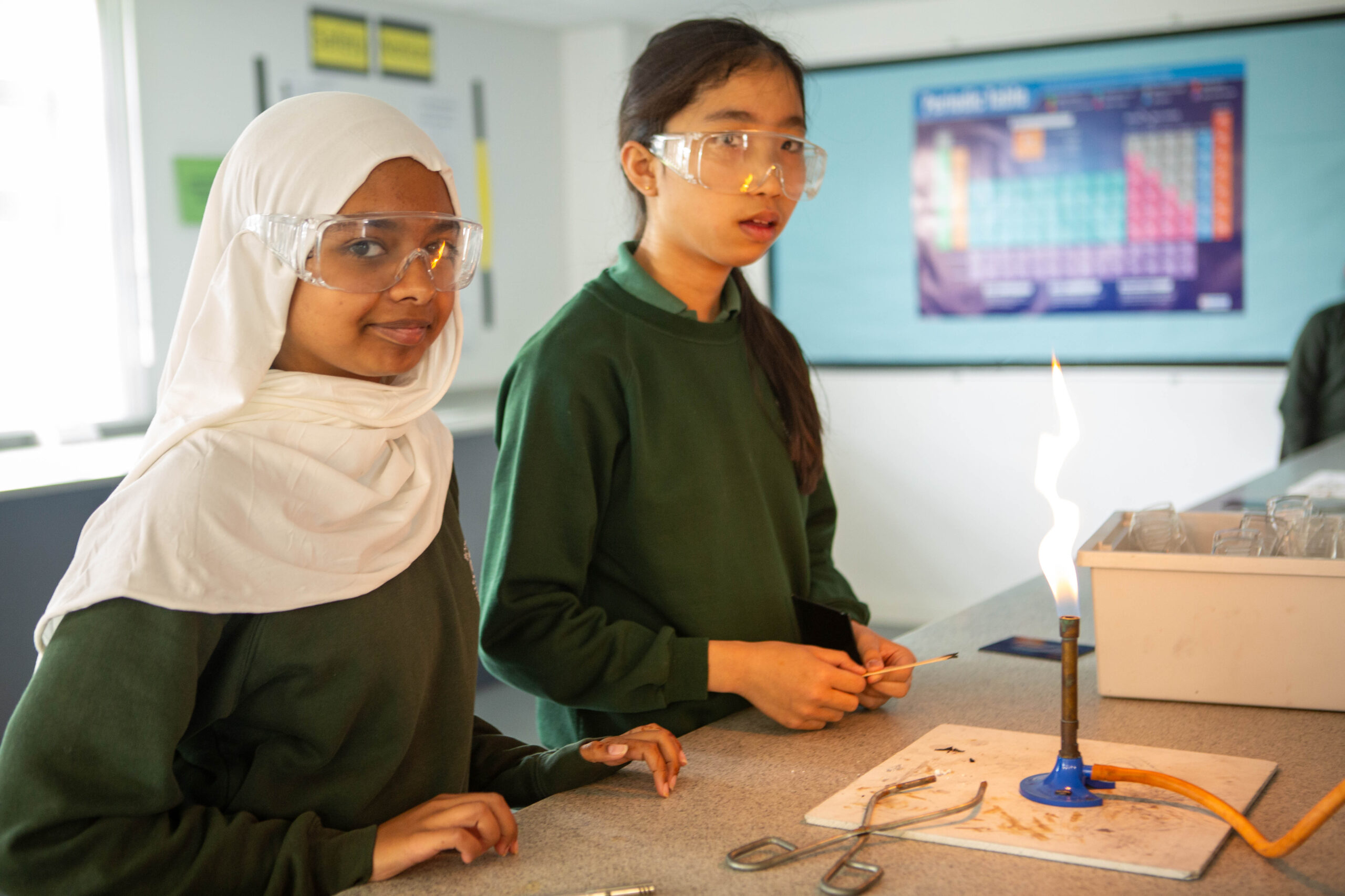 Two pupils are standing at a desk. One pupil is wearing a white headscarf and protective eyewear. The other pupil is wearing a green jumper and protective glasses. The table has a lit Bunsen burner.