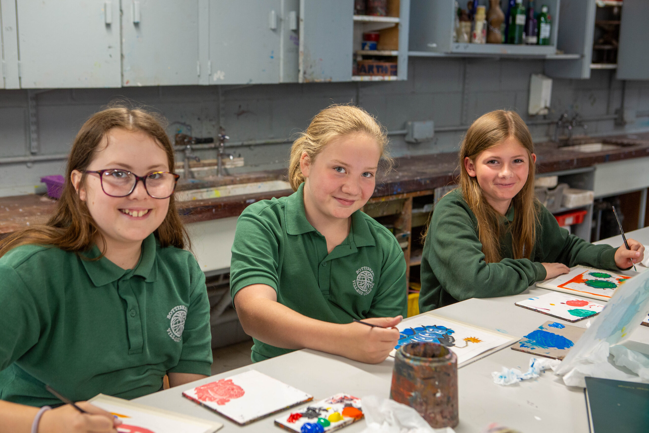 Three pupils are sitting at a desk. They are smiling at the camera. They are wearing green jumpers.