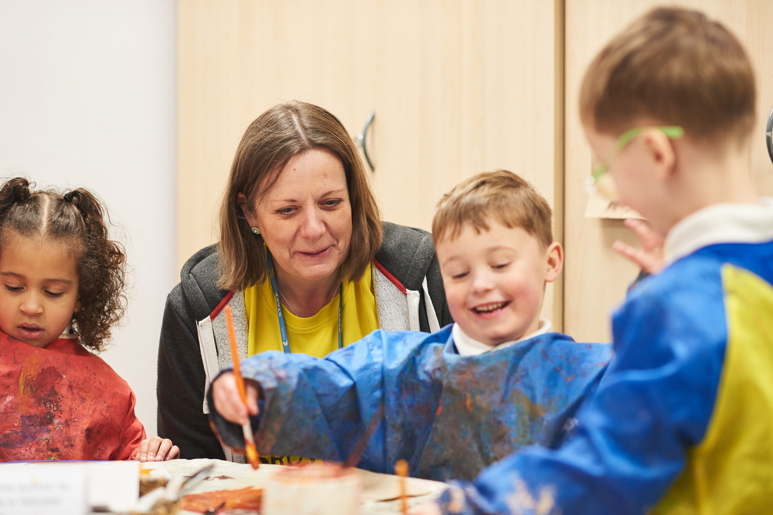 Three pupils are sitting at a desk with a teacher. They are wearing waterproof bids and painting on pieces of paper.