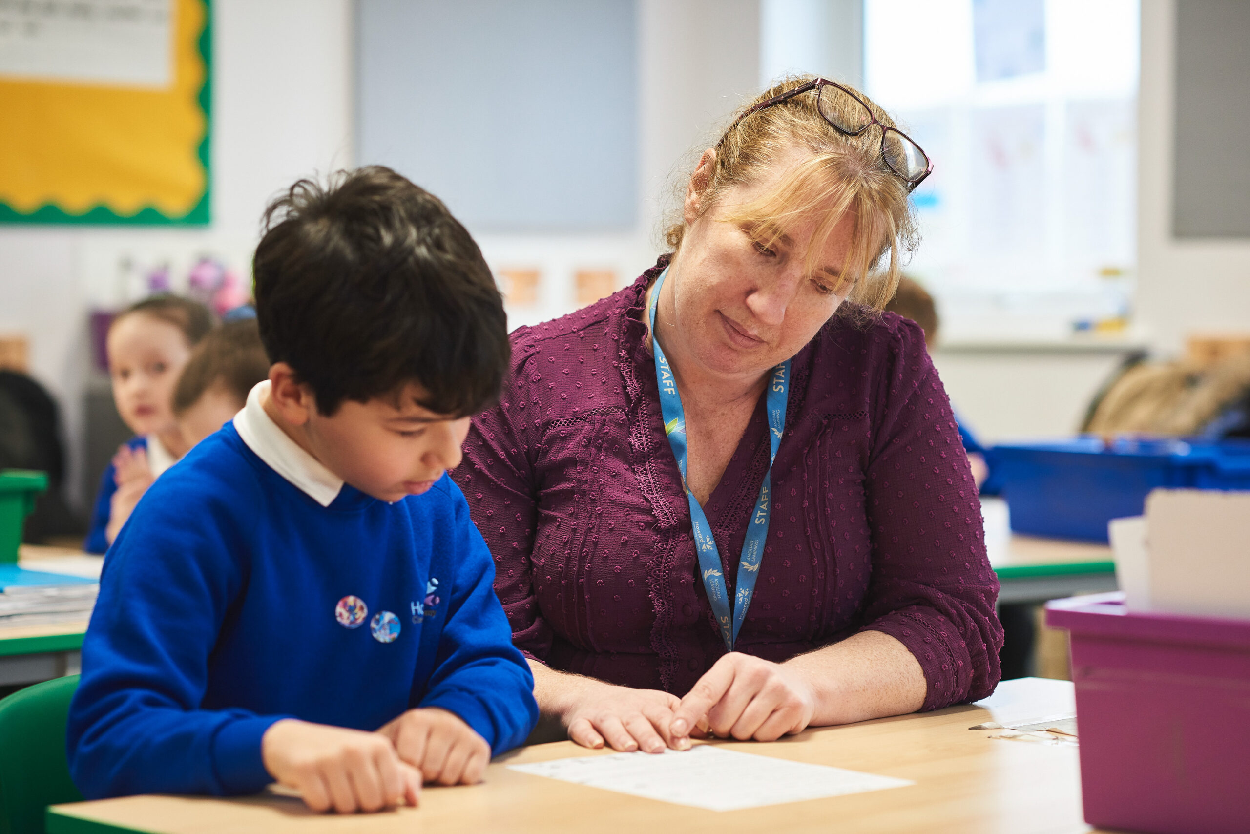 A pupil is sitting at a desk with a teacher. They are reading from a piece of paper. The pupil is wearing a dark blue jumper, the teacher is wearing a purple top.