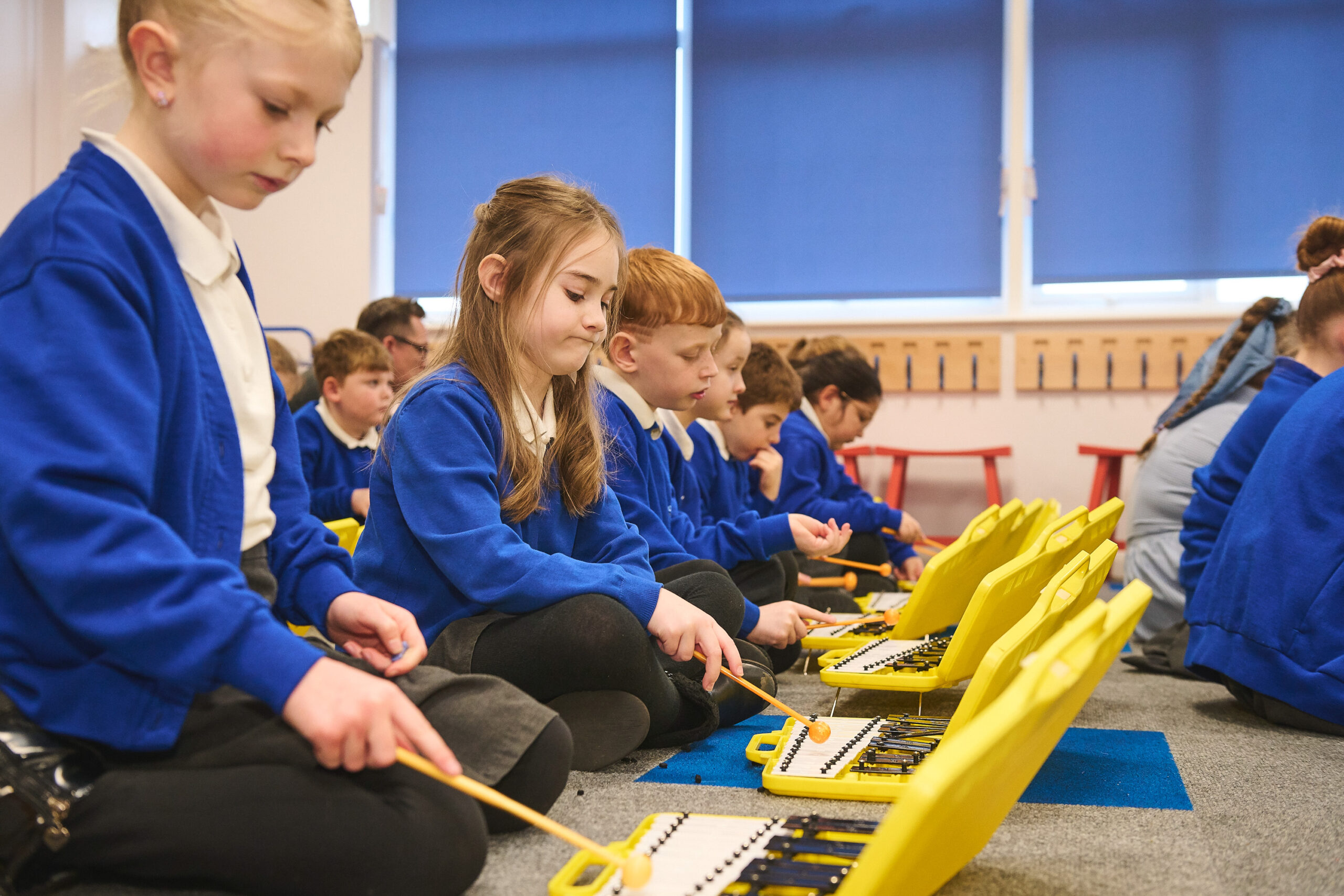 Pupils are sitting in a line on the floor. They are playing on a yellow Xylophone.