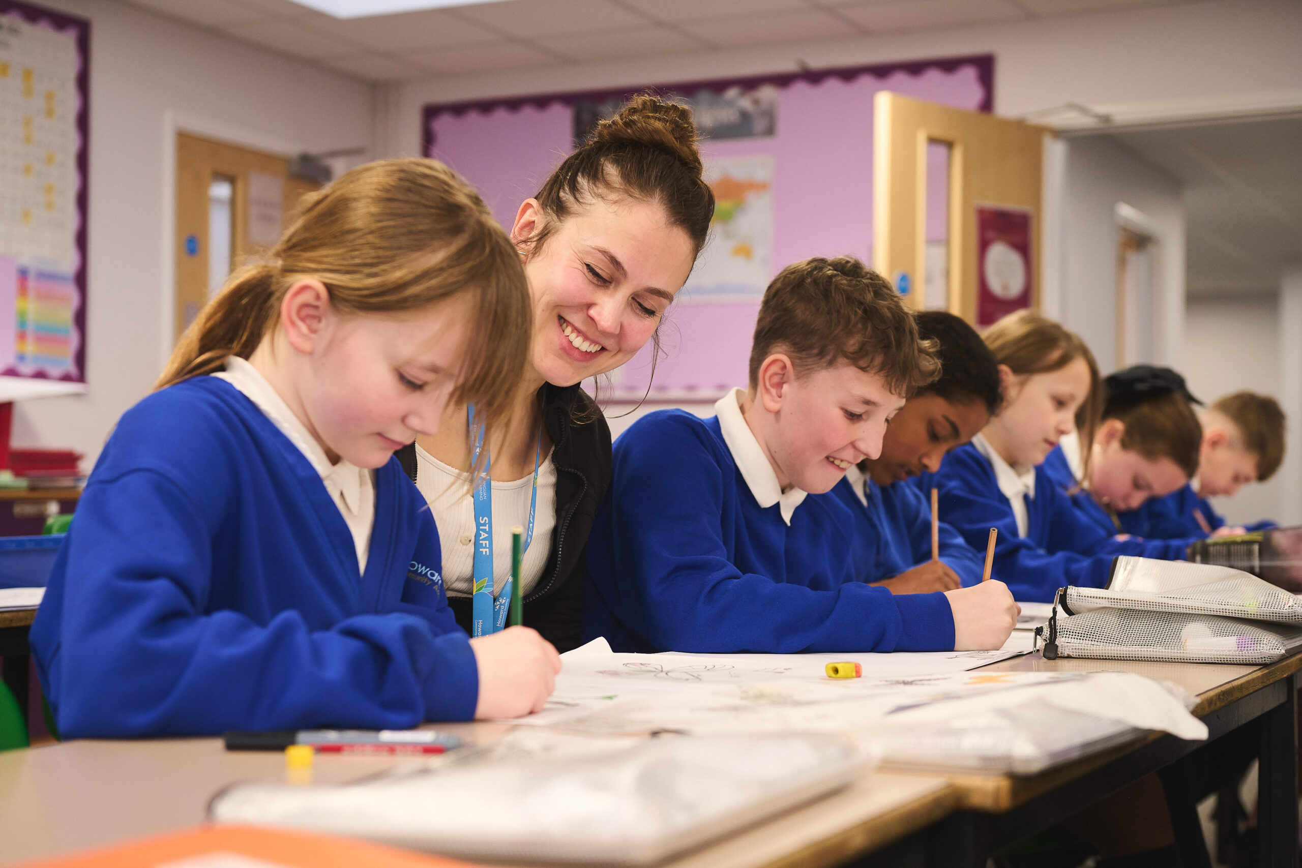 Pupils are sitting at a desk in a line. A teacher is sat in between two pupils. The teacher is smiling at the pupil, who is writing in their book.