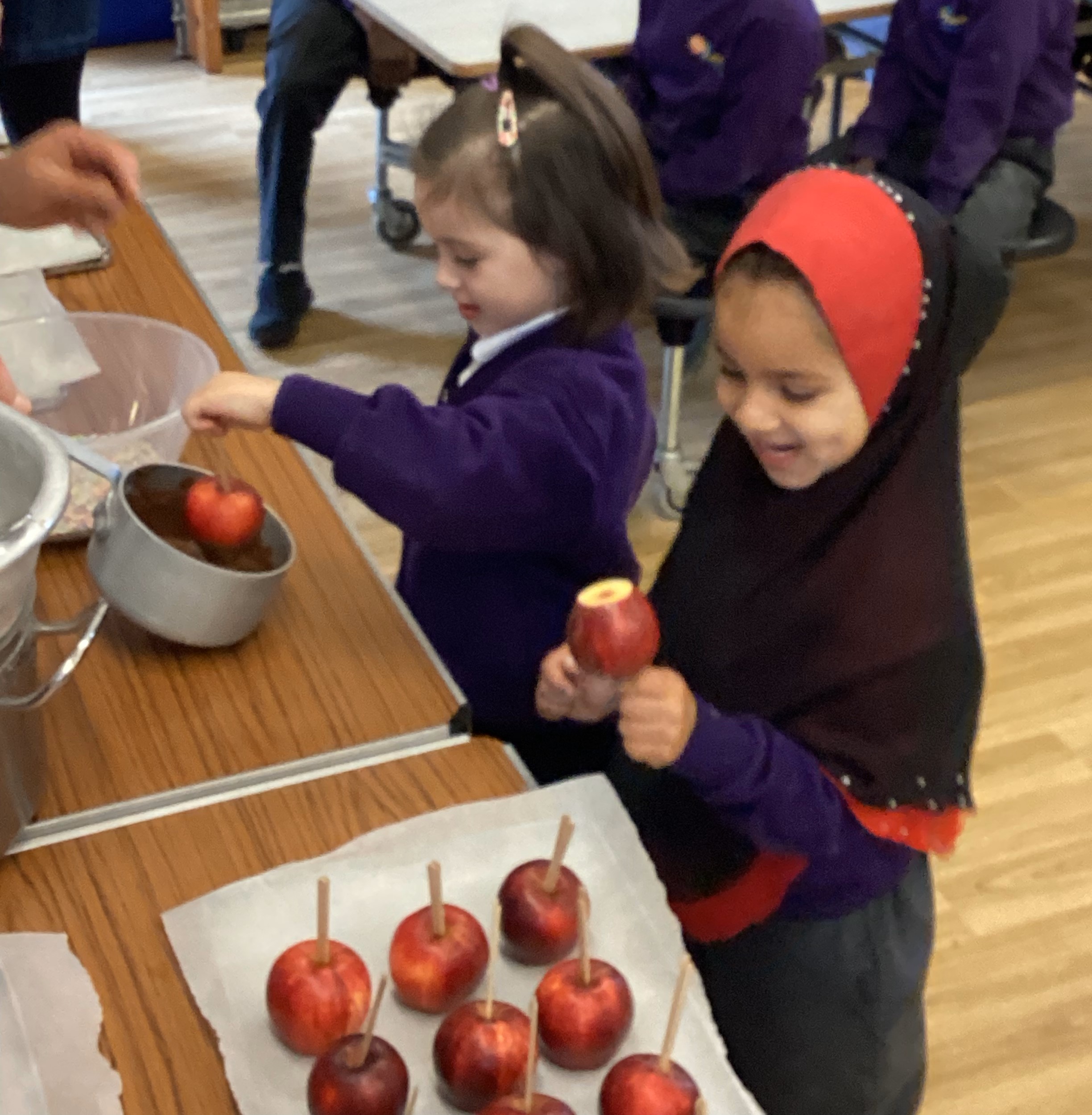Two pupils, one wearing a purple jumper and the other a black and orange headscarf, are placing sticks into apples and dipping them in chocolate.