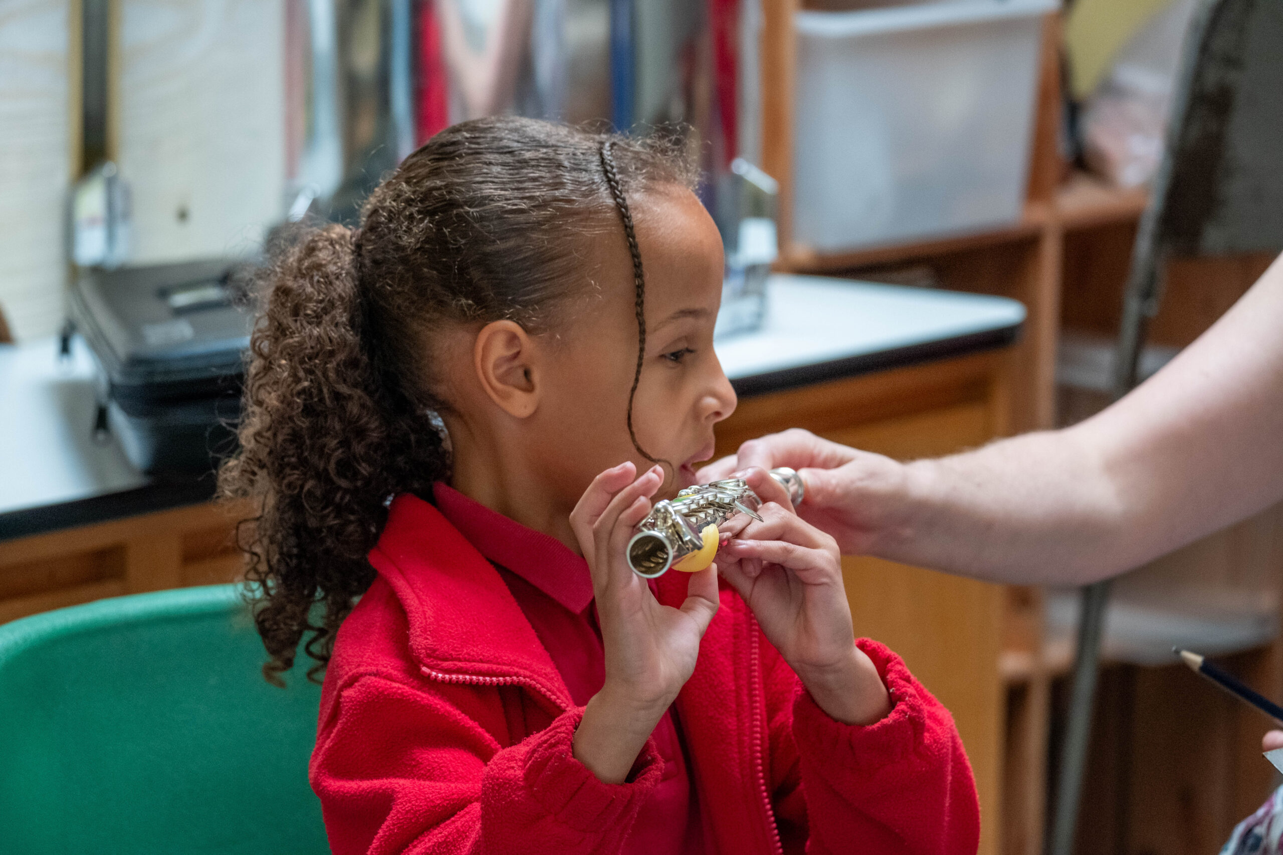 A young girl is playing the flute, looking away from the camera. She is wearing a red polo and red jumper.