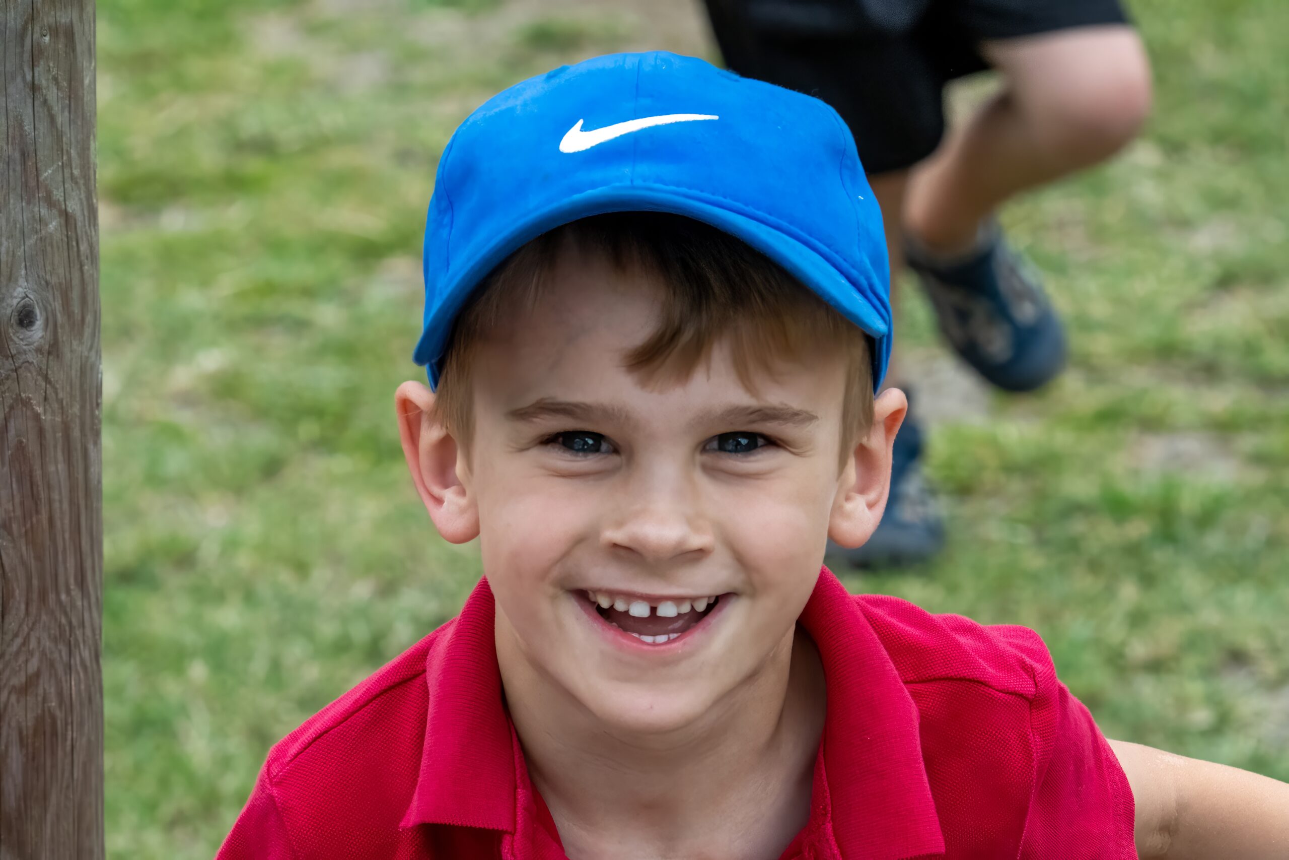 A young boy is smiling at the camera. He is wearing a blue cap and red polo.