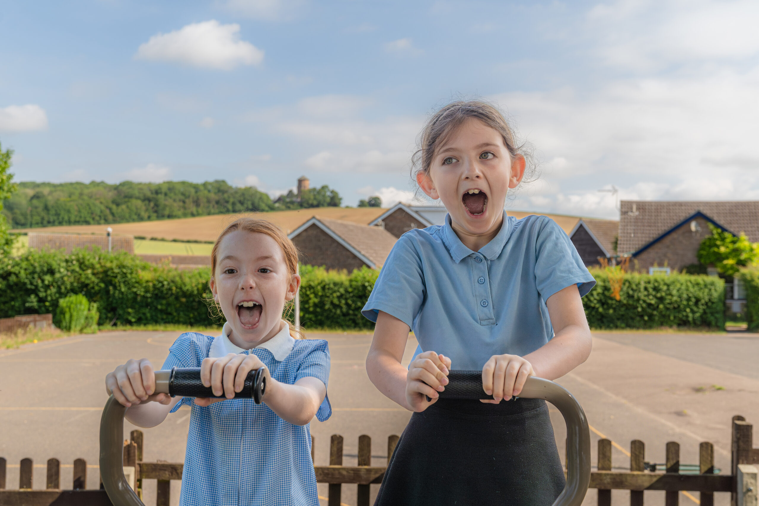 Two pupils are standing on a piece of aparatus with a field behind them. They have their mouths open at the camera.