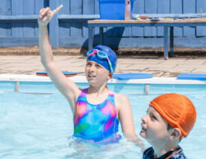 Two pupils are in a swimming pool. One pupil is wearing a blue cap with goggles. Another is wearing an orange swim cap.