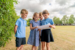 Four pupils are standing in a field. Two of the pupils are holding a piece of paper. One other pupil is staring at the paper with his hands behind his back.