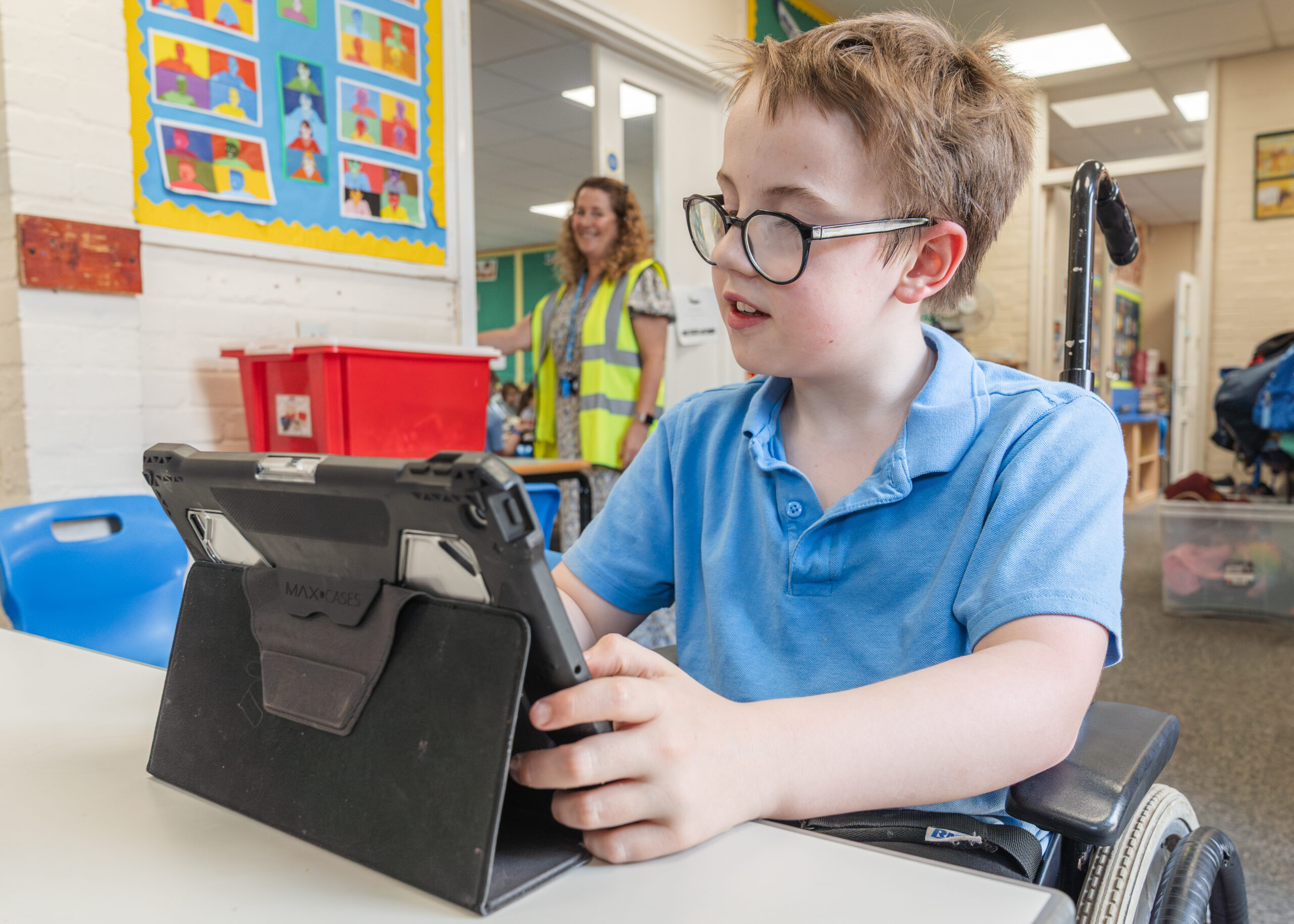 A pupil in a wheelchair is sitting at a desk and holding an iPad. He is wearing glasses and a blue polo shirt.