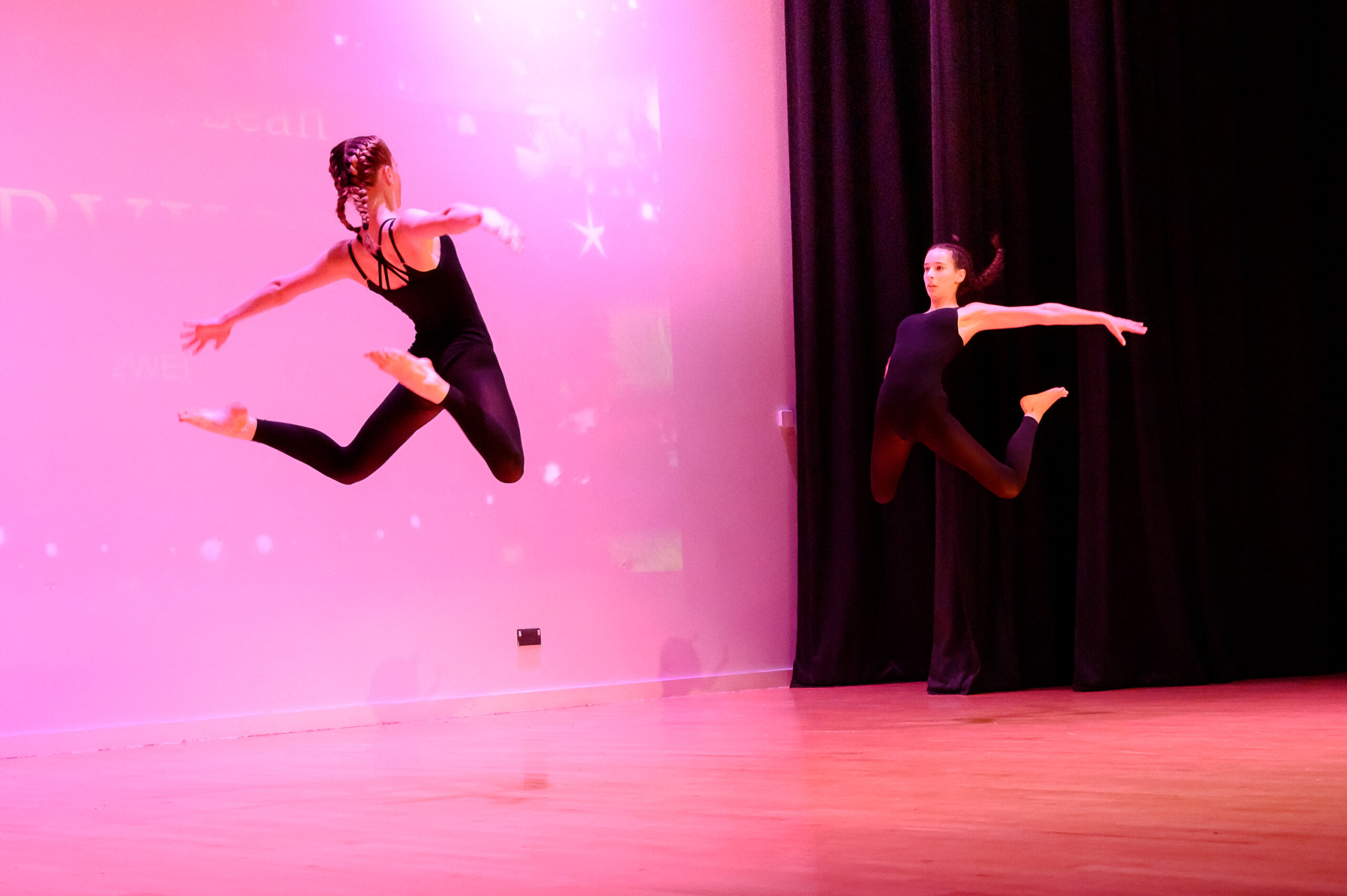 Two pupils are performing on stage in black leotards. They are facing eachother and leaping in the air.